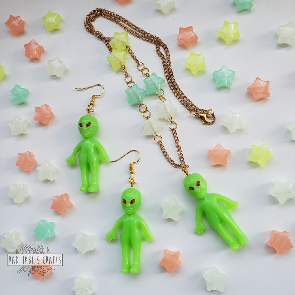 A matching set of green alien jewellery, two earrings and a necklace, on a white background surrounded by pink, blue, white, and green stars.