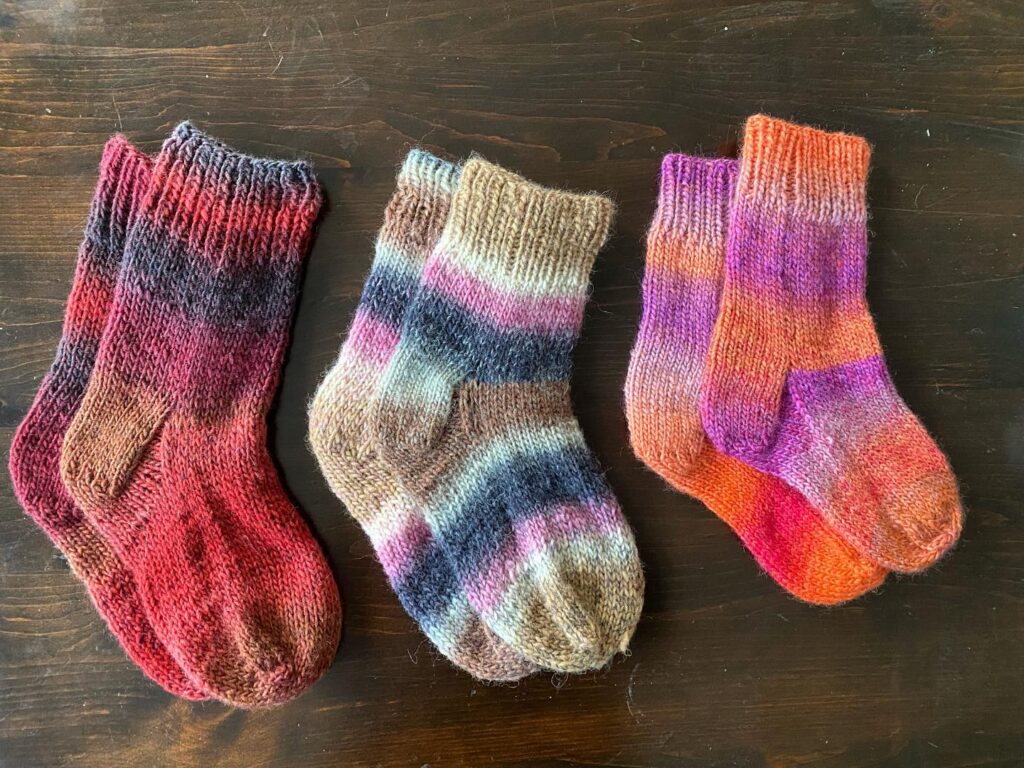Three pairs of striped wool socks in various sizes. One pair is red and brown, the second is brown, blue, and pink, and the third is orange, pink, and purple. They are lying on a dark brown wooden background.