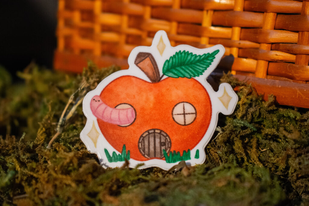 A sticker of a red apple with 2 circular windows, a brown wooden door, and a pink worm with rosy cheeks that is smiling. The apple has a big green leaf and sparkles. Behind the sticker is a pile of green moss and a wicker basket.