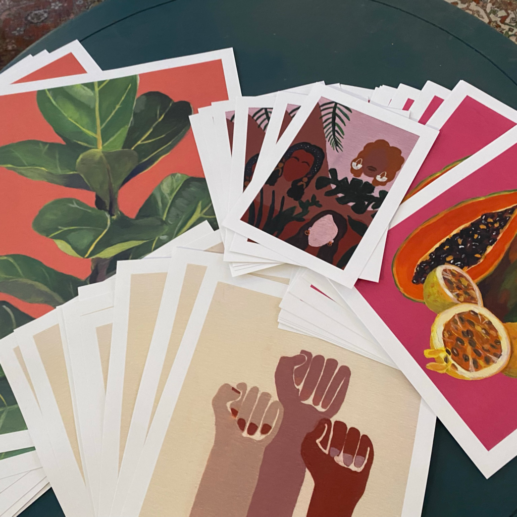 Stacks of multiple-sized prints layered on a teal table. The prints show a fiddle leaf fig with a bright coral background, a small print of 3 women's faces with plants and pink-toned background, a papaya cut in half and guava halves with a bright pink background, and 3 fists with painted nails on a beige background.